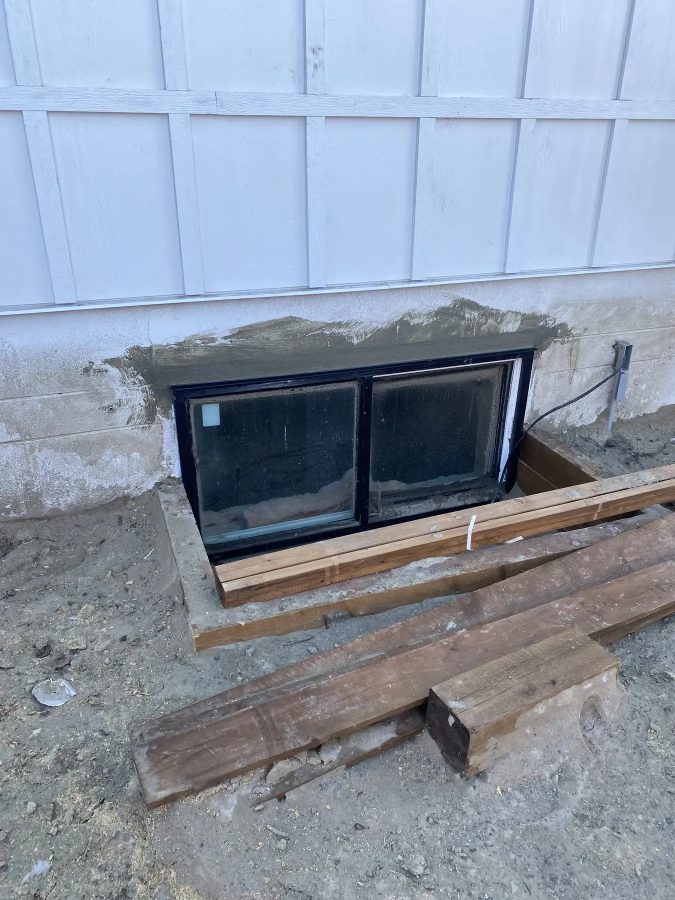 Enlarged basement window opening in a concrete foundation wall with fresh parging around the new black frame and timbers stacked beside the well