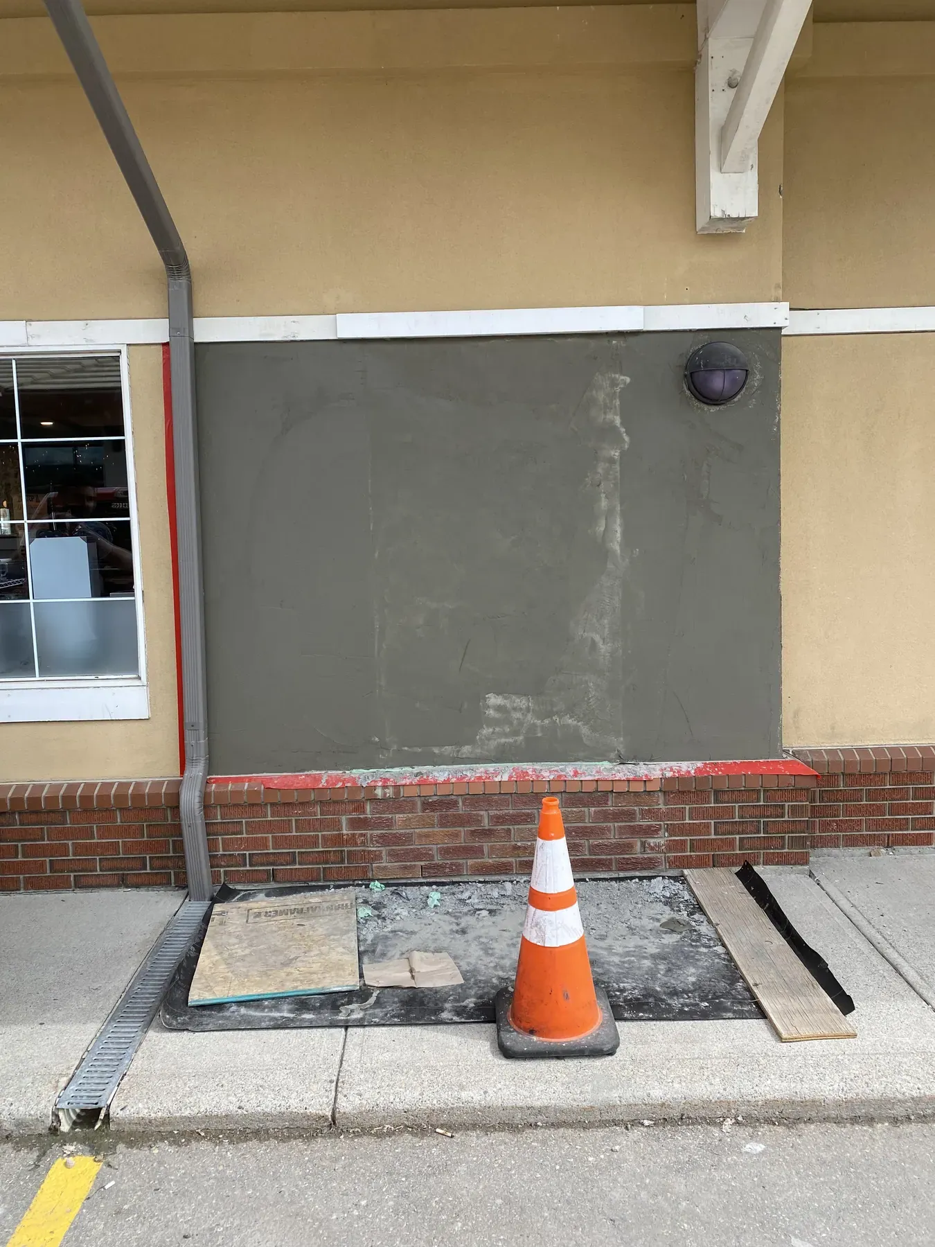 Grey stucco patch repair on a retail wall above a brick base, with a safety cone on the sidewalk
