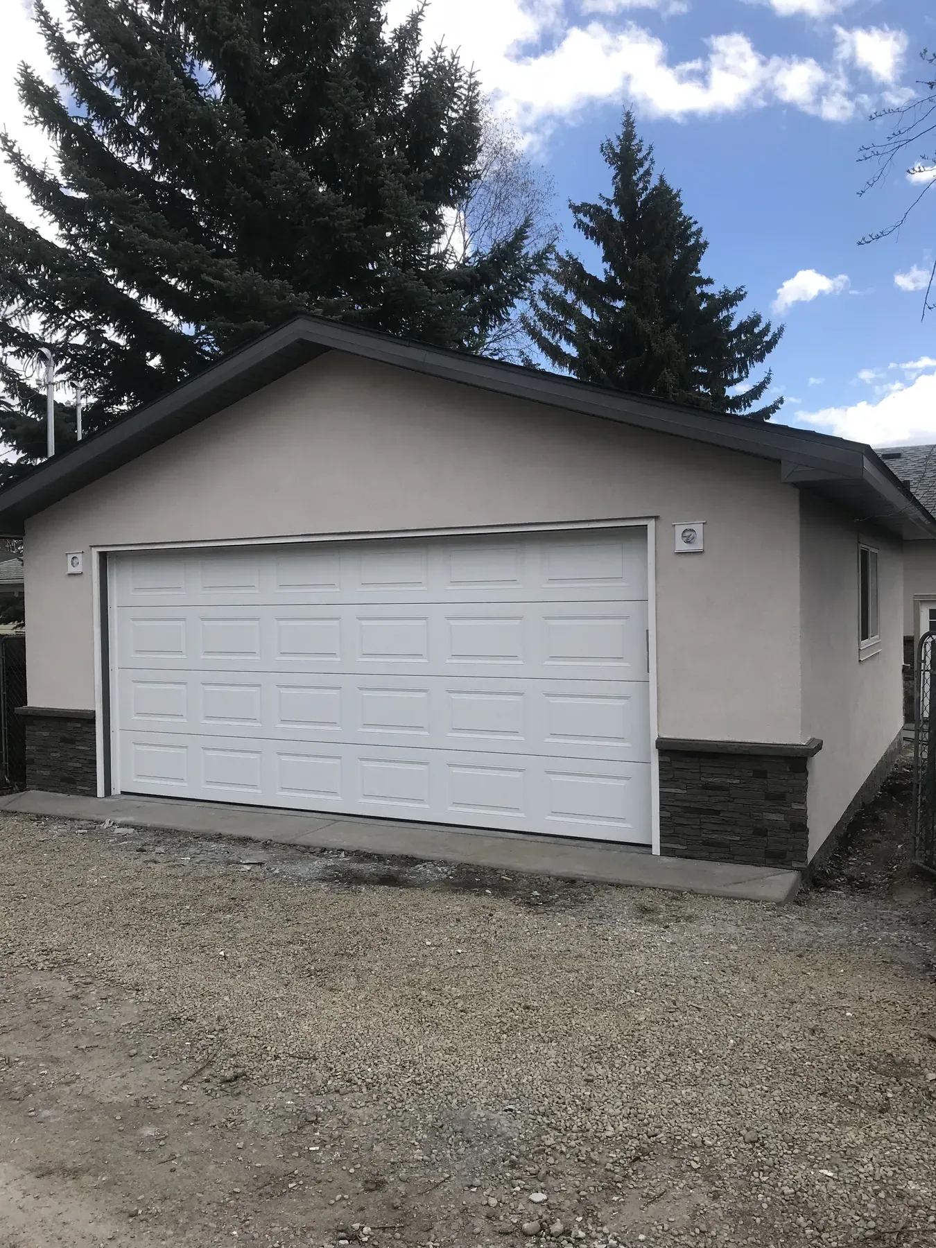 Finished detached garage with beige stucco, dark stone skirting and a white overhead door