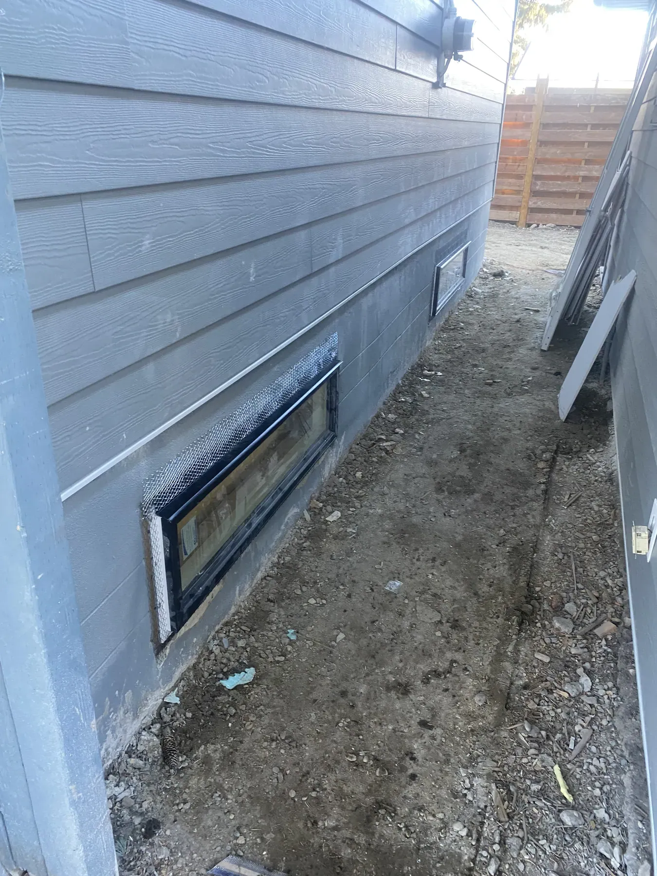 Grey fibre cement lap siding in a narrow side yard, with metal lath framing a basement window for parging