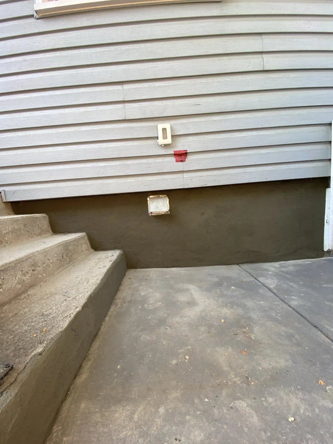 Fresh cement parging on a foundation wall beside concrete front steps, below grey vinyl siding