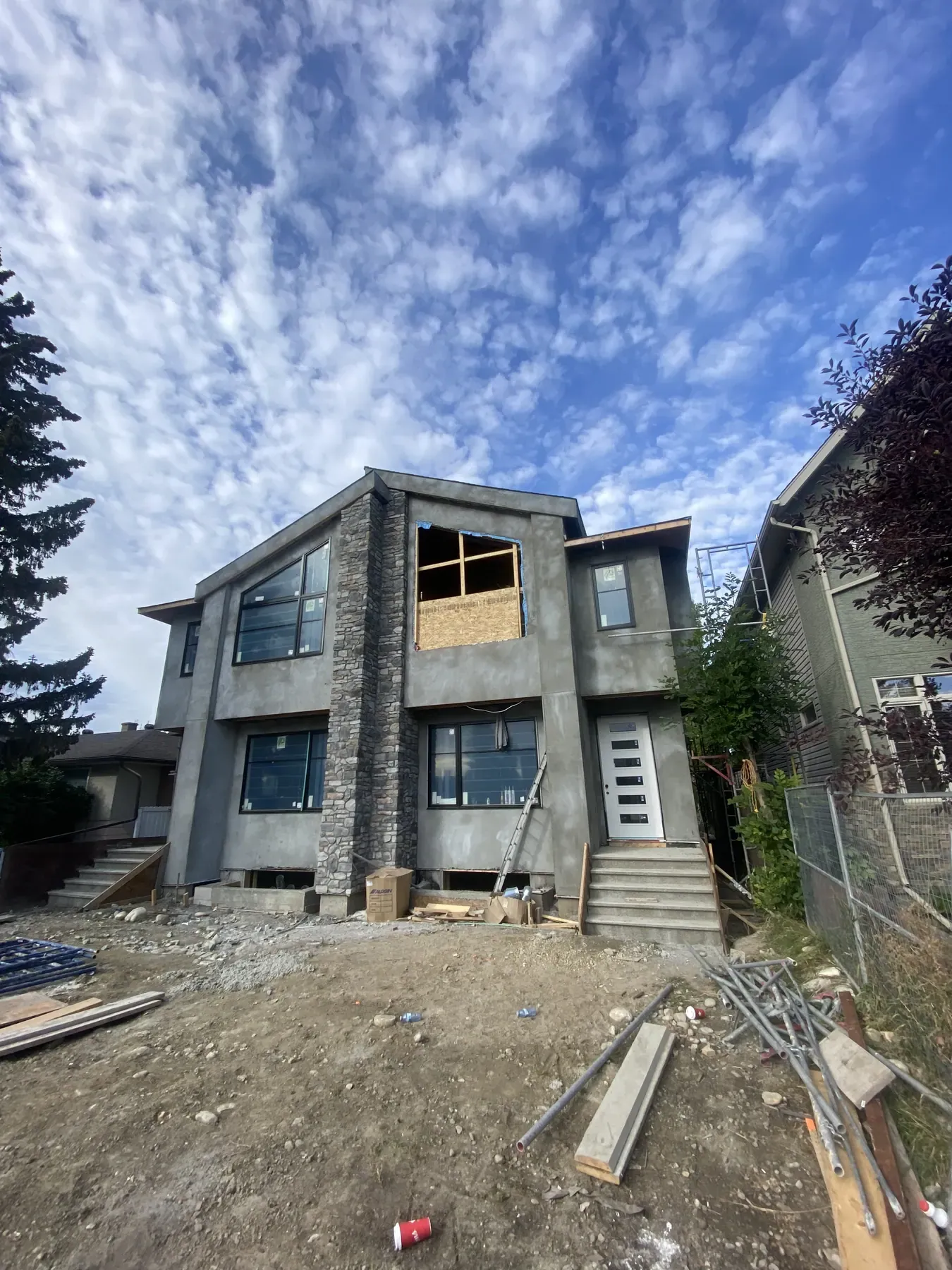 New two-storey infill home under construction with a grey stucco base coat, a stone veneer column and window openings boarded up