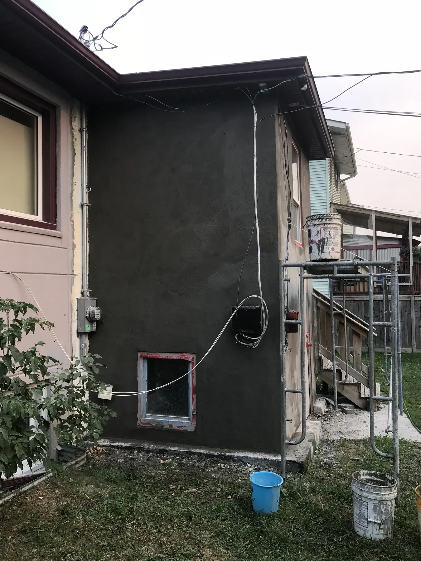 Side wall of a house covered in a fresh dark grey brown coat, with a taped basement window and scaffolding