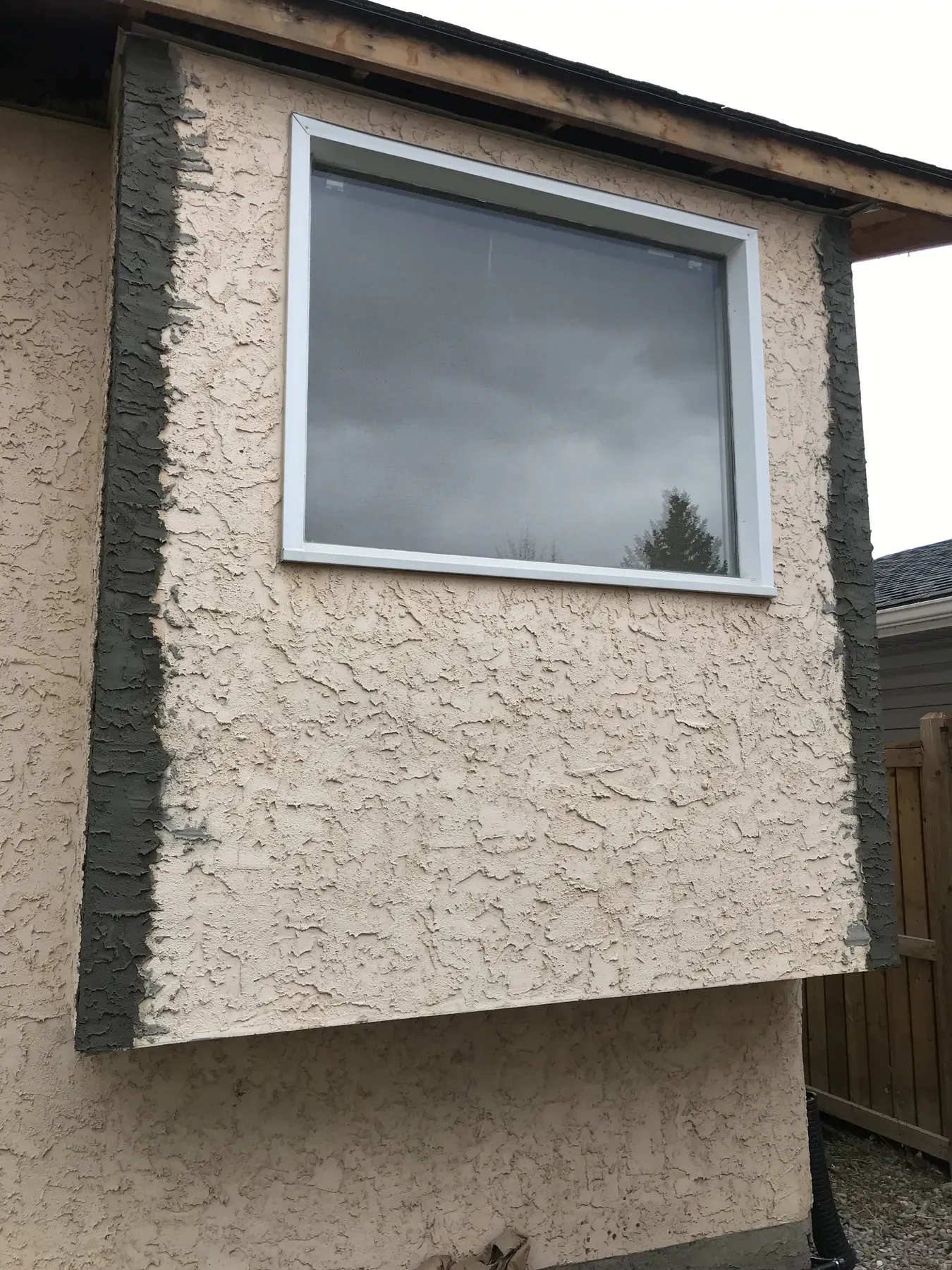 Beige textured stucco on a window bump-out with grey base coat patches at both edges, before the finish colour match