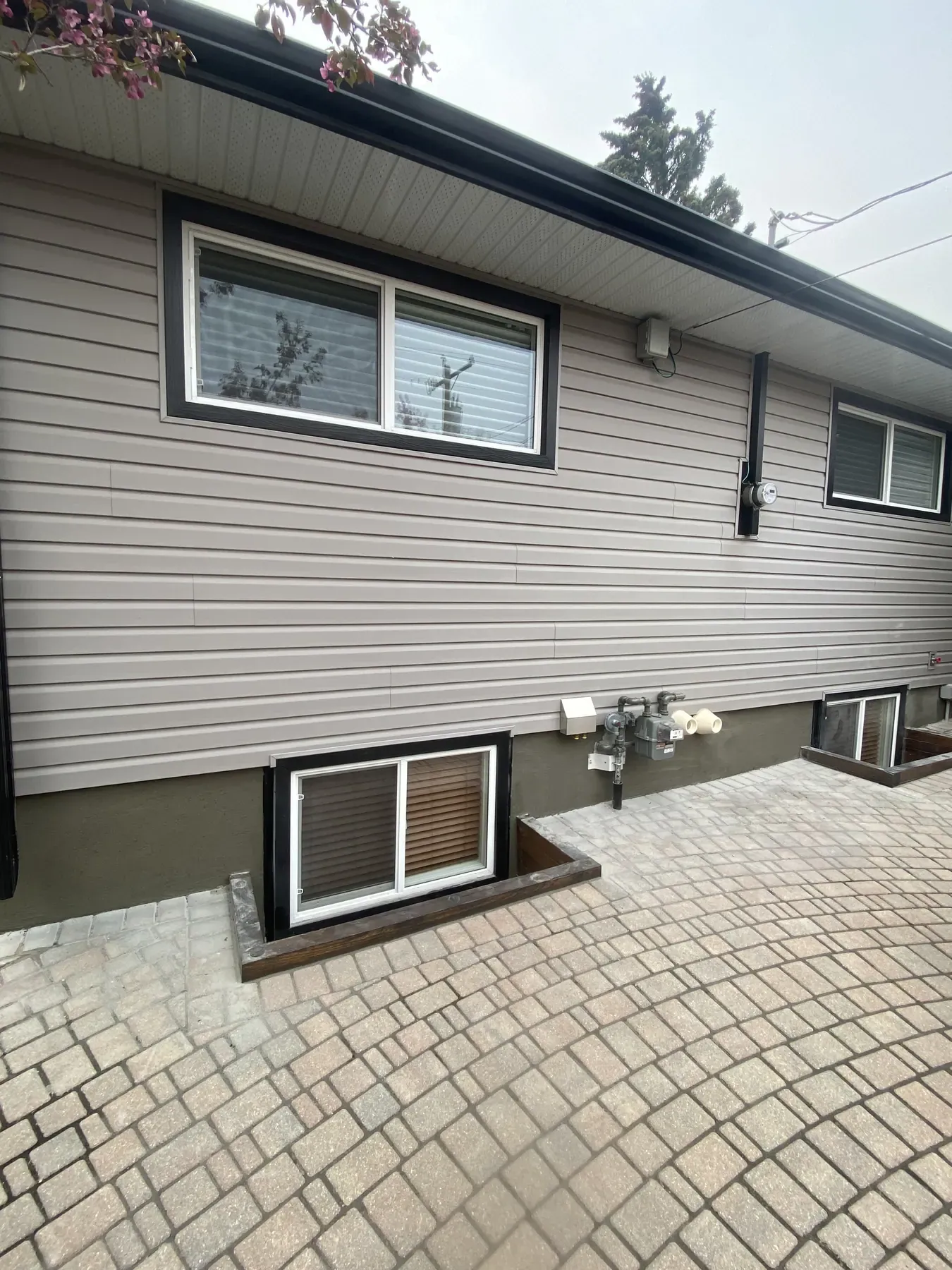 Tan vinyl siding with black-trimmed windows above a parged foundation and basement window wells on a paver patio